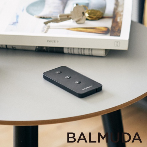 黑色遙控器，設有電源、風速調整和擺頭按鈕，以及BALMUDA品牌標誌，放置於白色圓形桌面。