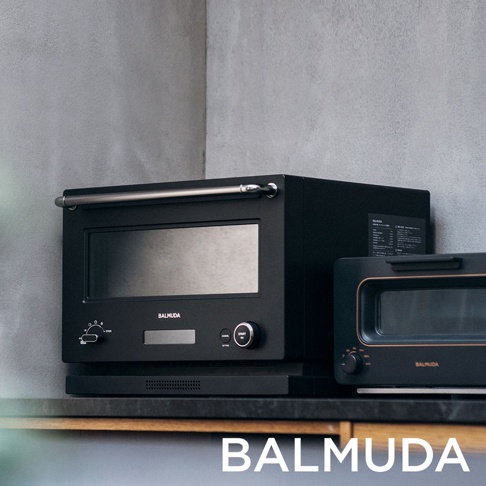 黑色 BALMUDA 微波烤箱，配有銀色提把、黑色玻璃門和數位顯示螢幕，左側有旋鈕和按鈕，右側另有一個黑色烤麵包機。