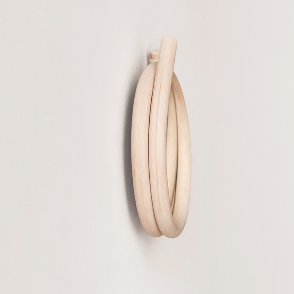 淺木色藤編圓鏡，鏡面為圓形，藤編外框呈彎曲環狀，掛在白色牆壁上。