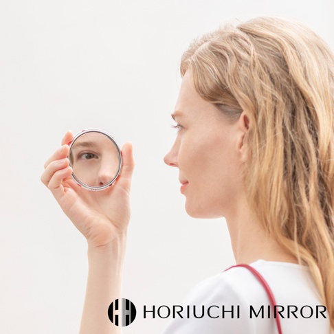 一位金髮女子手持一個約 7 公分直徑的銀色金屬圓形化妝鏡，鏡中映照出她半張臉。