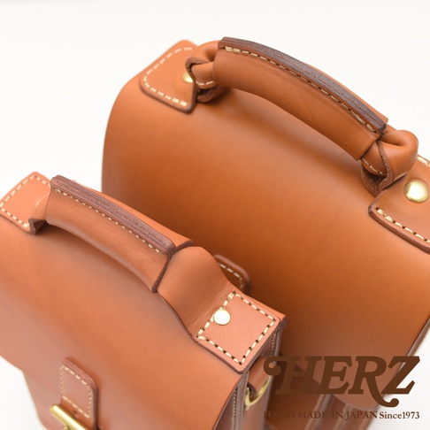 兩款職人手作皮革單肩包，顏色為棕褐色，材質為真皮，帶有經典手縫線與金屬扣件，設計簡約實用。