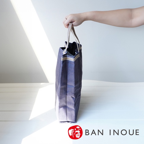 一個深藍色棉織隨身購物袋，提把為條紋織帶，袋口有裝飾性滾邊，由人手提起。