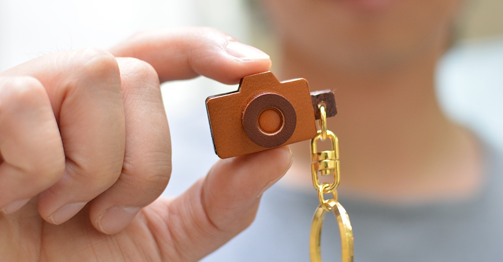 職人手作皮革相機造型鑰匙圈，咖啡色皮革製成，鏡頭部分為深咖啡色皮革裝飾，搭配金色金屬掛環與鍊子。