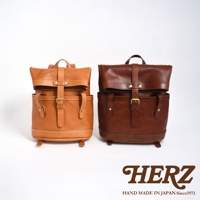 職人手作皮革雙口袋折蓋後背包，左為淺棕色，右為深棕色。兩款背包皆為皮質，設有雙前口袋、折蓋設計，並以皮帶及金屬扣環固定。