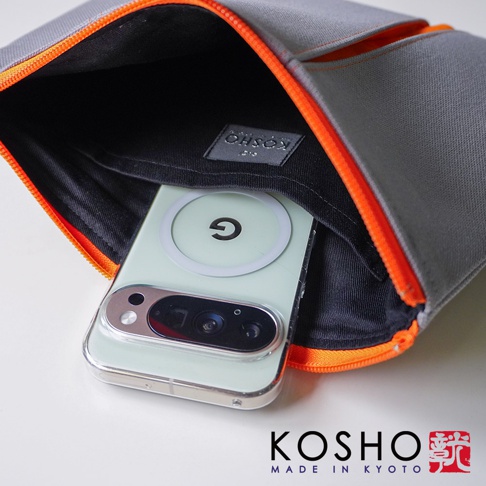 一個灰色的KOSHO品牌帆布收納包正半開著，內襯為黑色，袋內裝有一支淺綠色手機，手機背殼裝有白色圓形磁吸環。收納包邊緣搭配亮橘色拉鍊，整體呈現簡約的設計風格，右下角印有KOSHO MADE IN KYOTO及品牌圖章字樣。