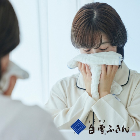 一位女性在鏡子前，用一塊白色、柔軟、多層次的洗澡巾擦拭臉部。洗澡巾邊緣帶有藍色滾邊。
