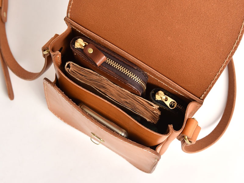 職人手作皮革翻蓋迷你單肩包，淺棕色，展示內部空間，可見一個筆記本、兩個拉鍊小包和一支筆。