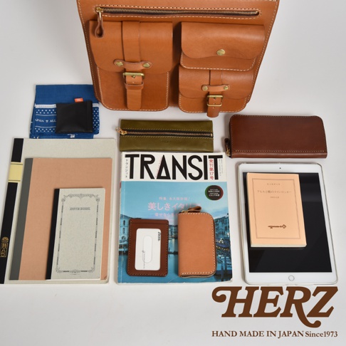 職人手作皮革三口袋三用後背包，展示多種搭配小物，包括筆記本、雜誌、證件套、鑰匙包、錢包及平板電腦。後背包為皮革材質，顏色為天然棕色，配有兩個前袋，袋蓋皆有金屬扣件及拉鍊，整體呈現簡約質感。