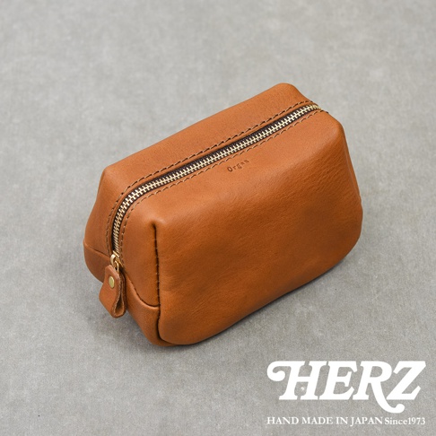 一個放置在灰色背景上的日本HERZ品牌Organ系列棕色皮革拉鍊收納包，皮革質感溫潤，包身側面印有細緻的Organ字樣，配有黃銅質感拉鍊，呈現簡約且經典的手作職人風格。