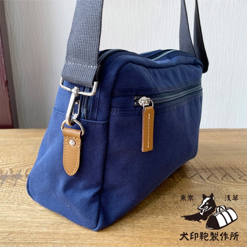 深藍色石洗帆布製成的斜背包，配有棕色皮革拉鍊吊飾和銀色金屬配件，右下角印有狗和行李的圖案。