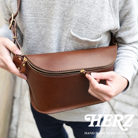 一個棕色皮革製成的立體斜背包，有金屬拉鍊和扣環，由一個人拿著。