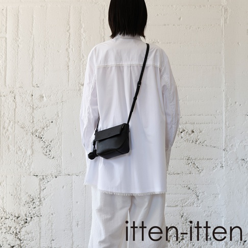itten-itten 品牌黑色皮革迷你單肩包，配有編織吊飾，由一位穿著白色襯衫和褲子的模特兒從後方展示。