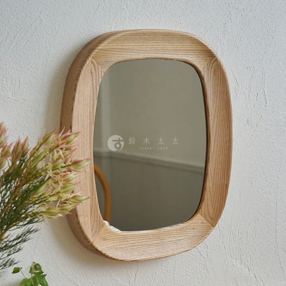 一個橢圓形、淺木色紋理的木框，內嵌一面鏡子，鏡框邊緣為圓潤的弧形，掛在白色紋理的牆面上，左側有綠色植物作為裝飾。