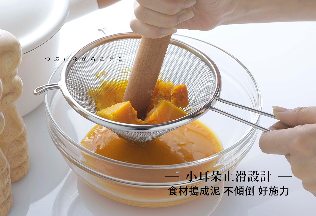 不鏽鋼錐形濾網，網孔細密，帶有兩個銀色金屬手柄，正用於將煮熟的南瓜塊壓成泥，南瓜泥與液體一同通過濾網流入下方的透明玻璃碗中。