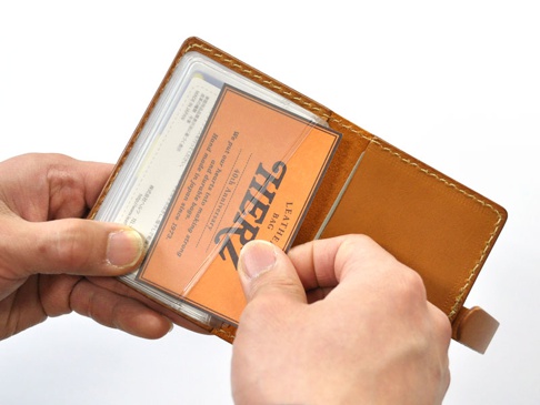 雙手持著一個棕色皮革製的卡片收納夾，夾內有數張卡片，其中一張橘色卡片印有黑色「HERZ LEATHER BAG」字樣，並有透明保護膜。