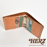 日本HERZ 職人手作皮革二折式薄型卡片鈔票夾，內有信用卡和鈔票，背景為白色，右下角有HERZ品牌標誌和 HAND MADE IN JAPAN Since1973 字樣。