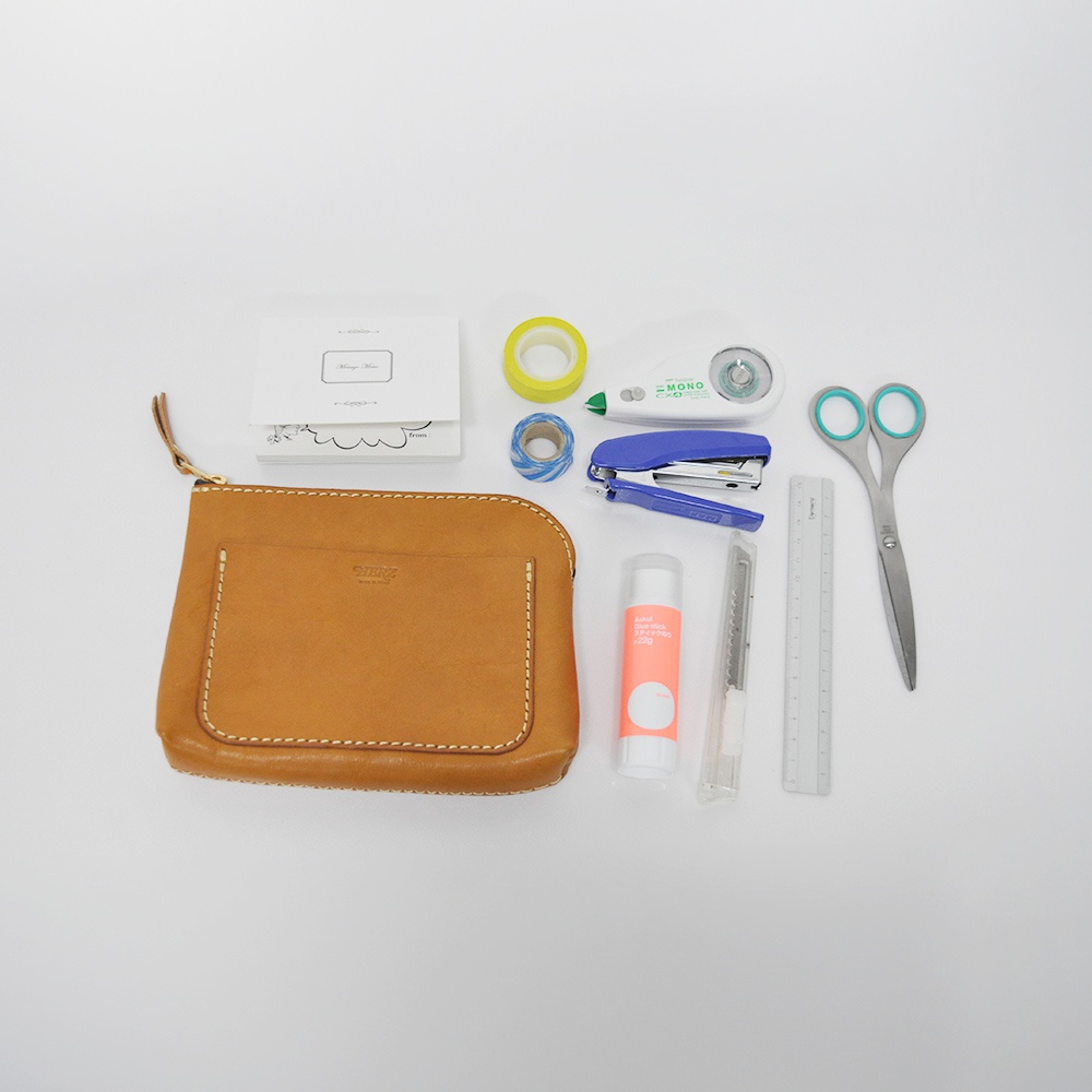 一個棕色皮革製的拉鍊收納包，正面有一個方形口袋，旁邊擺放著文具，包含藍色釘書機、白色修正帶、剪刀、尺、美工刀、膠水棒、黃色膠帶及藍白條紋麻繩。