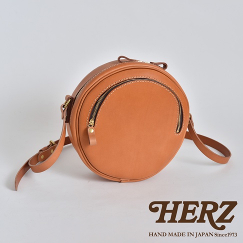 棕色皮革製成的手工圓形小包，飾有雙拉鍊開口和可調式背帶。