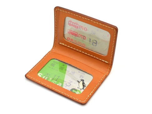 職人手作皮革雙折票券收納夾，呈現溫潤的棕褐色皮革，搭配清晰的縫線。內部設有兩個透明夾層，分別展示一張綠色Suica卡及一張PASMO卡，卡片上印有電車圖案與數字。