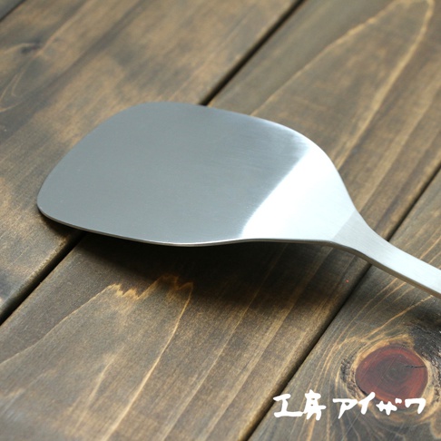 銀色不鏽鋼材質的萬用鍋鏟，鍋鏟頭呈寬大弧形，邊緣光滑，手柄部分延伸自鍋鏟頭，整體呈現金屬光澤，擺放在深棕色木紋背景上。