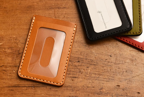 職人手作皮革直式票券收納夾，主體為淺棕色皮革，配有透明視窗，邊緣縫線為米白色。背景為木質桌面，另有黑色、軍綠色和紅色皮革收納夾部分入鏡。