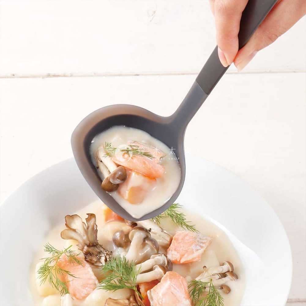 一手拿著marna TRYANGRIP三角耐熱矽膠料理湯杓，湯杓盛著一勺濃稠的奶油鮭魚燉湯，裡面有鮭魚塊、蘑菇和蒔蘿，湯勺的灰色和食物的白色、粉色與綠色形成鮮明對比，湯勺正從一個裝滿相同燉湯的白色盤子中舀起湯。