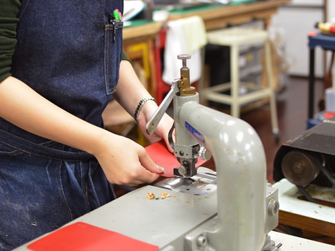 一名身穿丹寧圍裙的工匠，正使用皮革壓紋機處理一張紅色皮革。