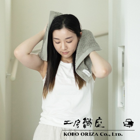 一位女性使用灰色人字紋有機棉毛巾擦拭頭髮，毛巾質感柔軟，品牌標籤清晰可見。