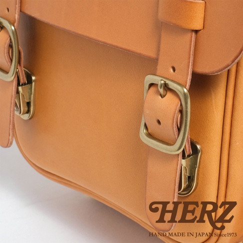 職人手作皮革小學生書包，淺棕色皮革，配有黃銅色金屬扣環和帶扣，設計簡潔，展現皮革的自然質感。