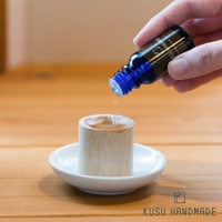 淺色木紋擴香木筒置於白色小碟上，一滴淺棕色液體從藍色蓋子的玻璃精油瓶滴落，木筒頂部有一個淺凹槽。