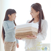 堆疊的純棉長毛巾，呈現白色、米色和淺棕色漸層。一位女士雙手捧著毛巾，一位小女孩站在旁邊。