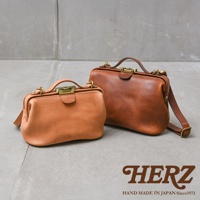 日本HERZ品牌，兩款不同尺寸的手工皮革Organ鎖孔式大開口肩背包，一淺棕色一深棕色，放置於水泥地上。