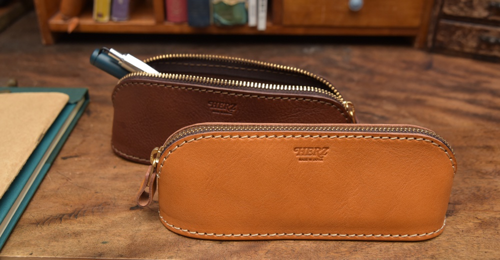 兩個職人手作皮革三角弧形收納筆袋，一個為深棕色，一個為淺棕色，均配有金色拉鍊和皮革拉環，表面壓印有品牌標誌。