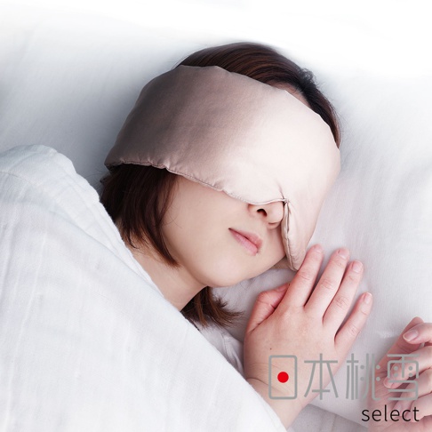 一位女性側躺在白色枕頭與床單上，配戴著 hiorie select 品牌的輕奢柔膚保濕絲綢眼罩，材質呈現細膩的香檳金色光澤，整體畫面營造出放鬆舒適的睡眠氛圍。