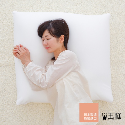 一位女子側臥在純白色大枕頭上，雙手交疊放在枕頭上，閉眼休息。枕頭材質看起來柔軟，表面光滑。背景為淺米色絨毛地毯。右下角有日文標示「日本製造 原裝進口」及品牌 Logo。
