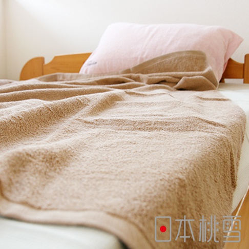 一張淺棕色超大浴巾覆蓋在床上，浴巾質地蓬鬆柔軟，局部有皺褶。床頭有一顆粉紅色枕頭，床架為木質。