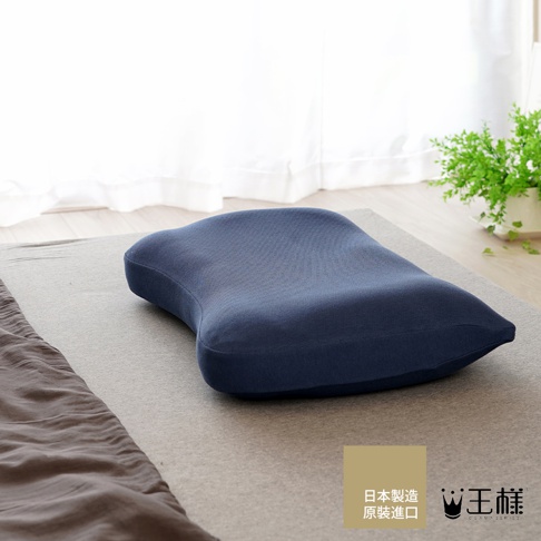 深藍色透氣材質的「王樣の極夢枕」男性專屬枕套，呈現人體工學的曲線設計，放置於灰色床墊上，背景為白色窗簾和綠色盆栽。