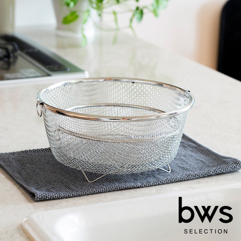 不鏽鋼圓形瀝水籃，網狀篩網設計，帶有兩個提把，放置在深灰色布墊上。