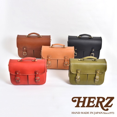 五款職人手作皮革單肩包，包含棕色、淺棕色、黑色、紅色和綠色。包款皆為兩用設計，附有提把與肩帶，並以金屬釦環裝飾。