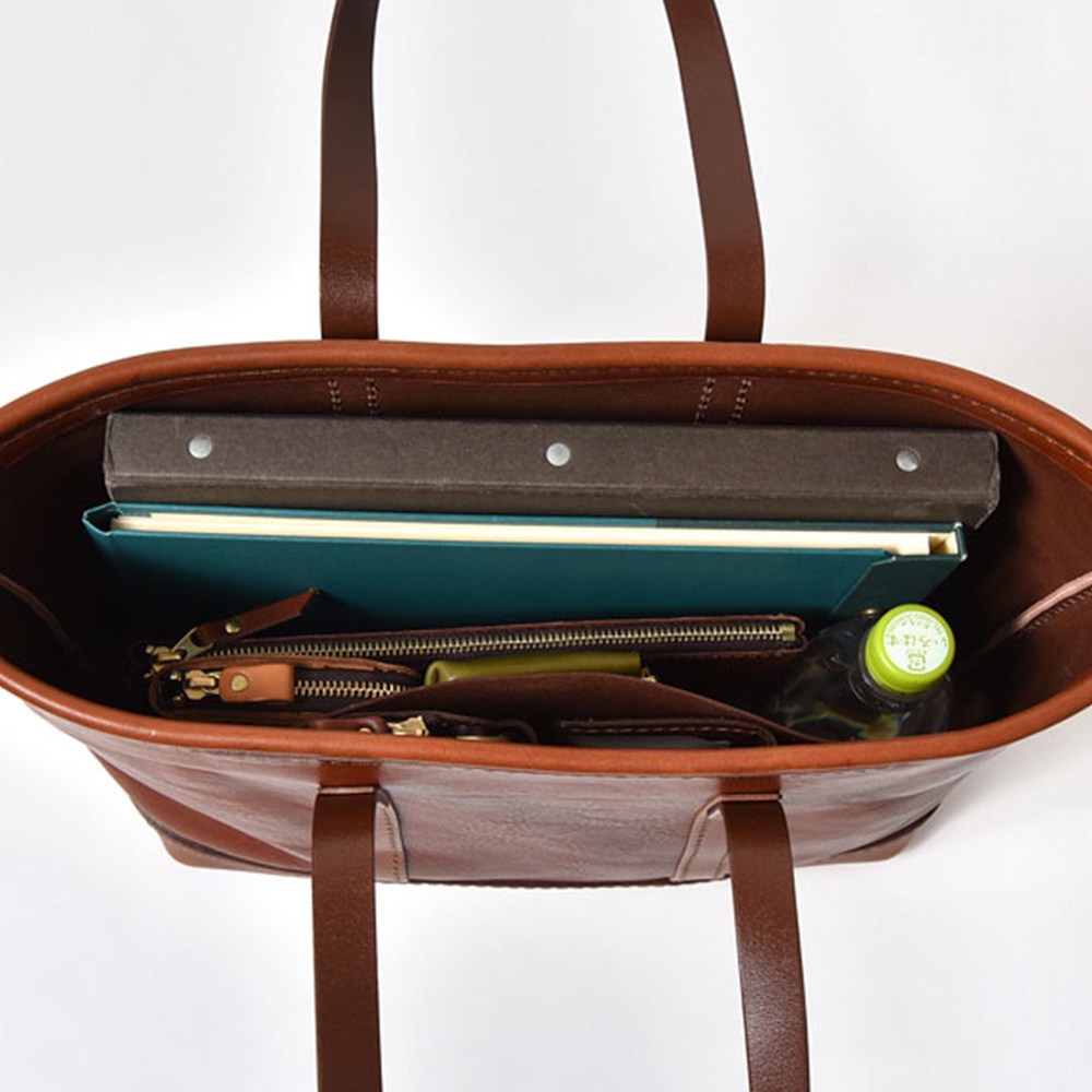 職人手作棕色皮革經典托特包，內部可見一本灰色筆記本、一本綠色筆記本、一個拉鍊零錢包、一個綠色長夾和一瓶水。