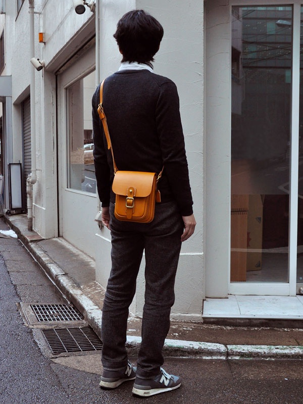 一位男士斜背著一個蜂蜜色皮革製的直式單肩包，包上有一個金屬扣和皮製的搭扣。