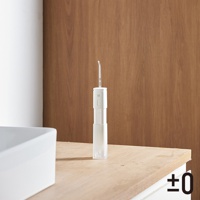 白色便攜沖牙機，機身上有 ±0 標誌與一個圓形按鈕。噴嘴為透明塑膠材質，向上延伸。產品放置於木質檯面上，背景為白色牆壁和木紋牆板。