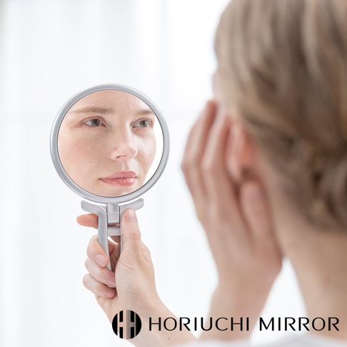 HORIUCHI MIRROR 品牌名稱以白色英文字母顯示在圖片底部，文字下方有一個圓形標誌，標誌由黑色線條組成，形狀類似於字母 H 的變形。畫面中央偏左，一只手正拿著一面銀色手持鏡，鏡中映照出一名女性的臉部特寫，女性眼神專注看著鏡子，臉部表情自然。鏡子邊緣為銀色金屬材質，鏡柄為可折疊設計，方便手持或立於平面。畫面的右側，可以看到另一名女性的側臉，她的頭髮為淺棕色，並用手輕觸臉頰。整體畫面背景為純