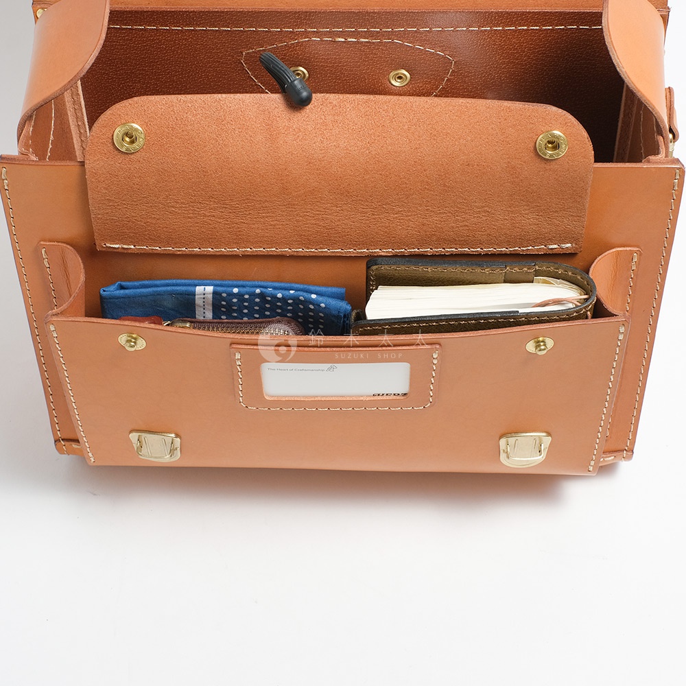 職人手作皮革橫式書包內部，展示著皮質的質感與精緻的縫線，中間隔層可放置藍色波點手帕與一本小筆記本。