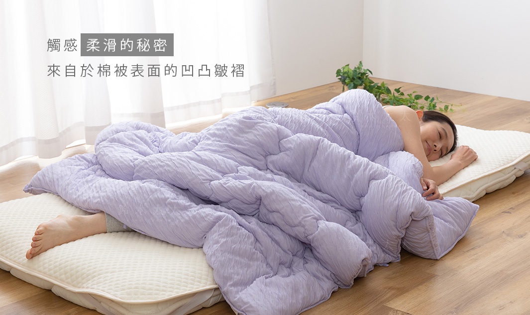 一床淺紫色舒芙蕾透氣四季被，表面有細緻的凹凸紋理，覆蓋在躺臥的女性身上，下方襯著米白色床墊。