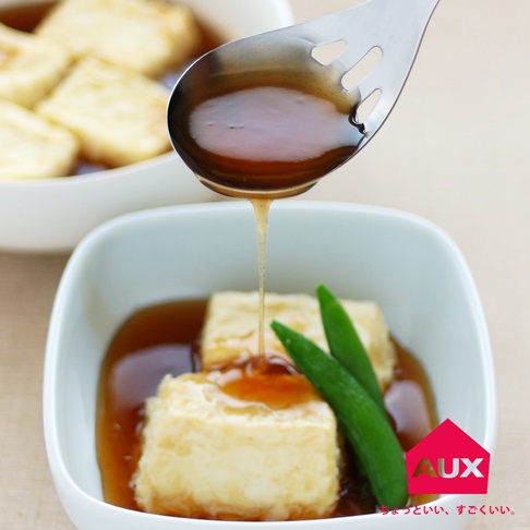 leye 濾水菜匙，銀色金屬材質，勺中盛有醬汁，正從菜匙中滴落，下方為裝有方形炸豆腐塊、淋上醬汁及綠色毛豆的白色方形碗。