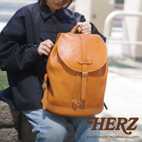 一位女士手持一個棕色皮革束口後背包，背包上有單扣和抽繩設計，並有白色縫線裝飾。