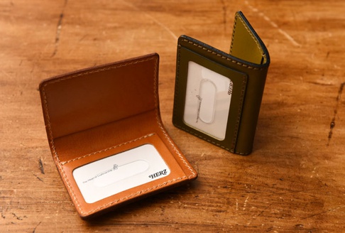 兩個職人手作皮革三面雙折票券收納夾，一個為棕色皮革，內有兩個卡槽，其中一個卡槽內放有白色卡片；另一個為橄欖綠皮革，有一個透明卡槽，內放有白色卡片。