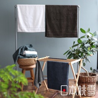 白色與深棕色浴巾掛在金屬架上，下方木架上有深藍色毛巾，籃子裡疊有綠色及白色毛巾，旁有綠色植物。