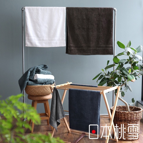 白色與深棕色浴巾掛在金屬架上，下方木架上有深藍色毛巾，籃子裡疊有綠色及白色毛巾，旁有綠色植物。
